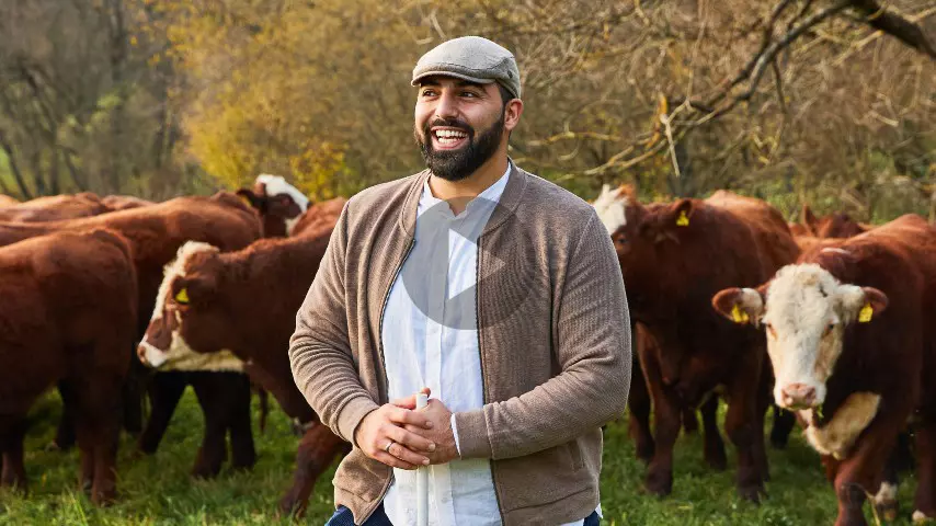 Josef steht lächelnd auf der Weide mit einer Herde Biorindern hinter ihm. Auf der Weide der Bio-Bauernhöfe